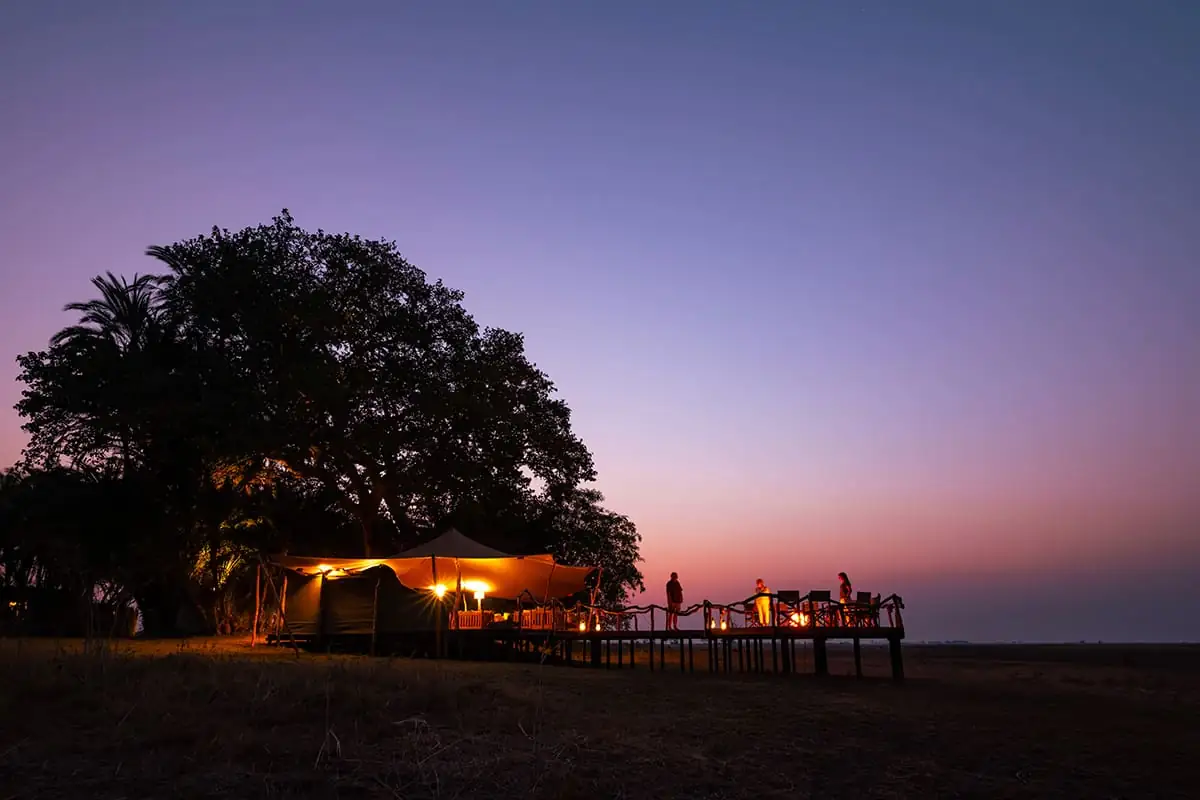 A lit tented camp on an open plain under a deep violet dusk sky