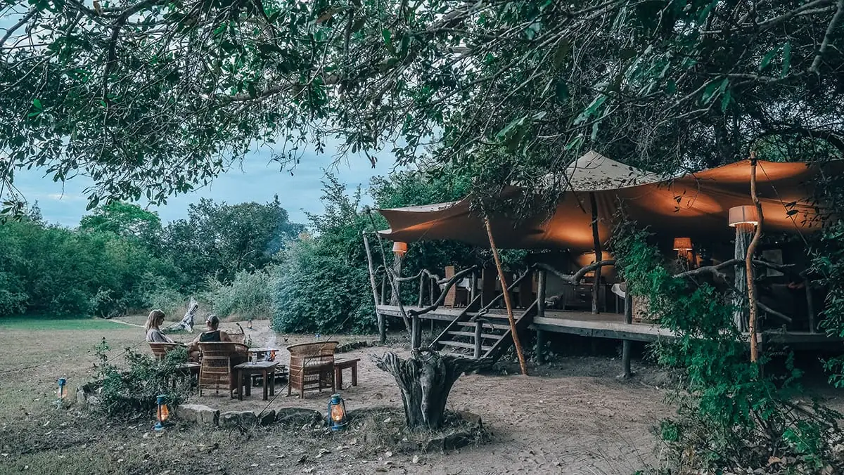 Guests gathered around a fire pit outside a tented camp at dusk
