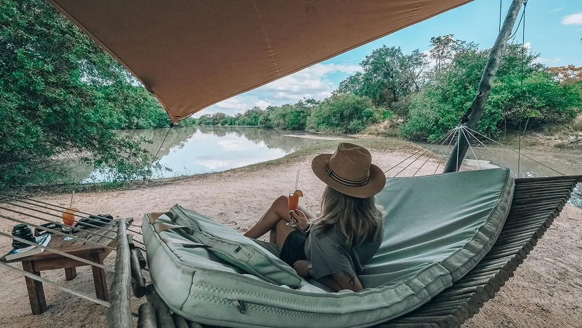 A guest resting in a hammock under a canvas awning, looking out over a waterhole